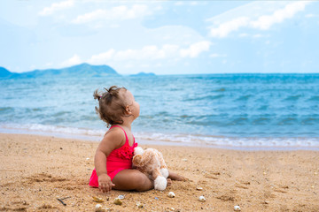 pensive little girl hugging teddy bear and looking away while sitting on seashore.