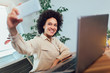 © Mediteraneo - Smiling young African female entrepreneur sitting at a desk in her home office make selfie photo.