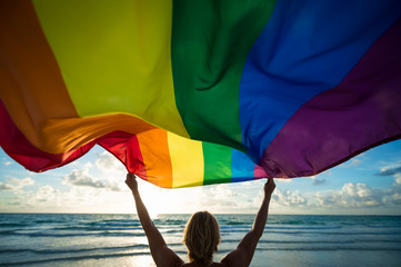 Colorful silhouette of a man with blond hair holding a gay pride rainbow flag blowing in the wind on a tropical beach with golden sun