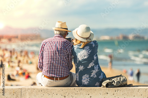 Cuadros en Lienzo Mature couple of retired lovers enjoying retirement on the beach facing the sea with mobile cell phone taking pictures at sunset