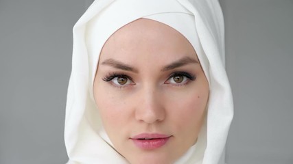 Wall Mural - Portrait of young attractive muslim woman wearing white hijab is rising her eyes, looking at the camera and smiling on light background.