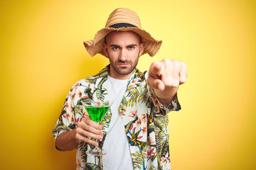 Wall Mural - Young man wearing summer hawaiian flowers shirt and drinking a cocktail over yellow background pointing with finger to the camera and to you, hand sign, positive and confident gesture from the front