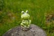 A frog on a rock sculpture beside a pond in the trees in a park.Decorative frog, statue, figure in garden.ceramic statue of a green frog.One of the variants of decor garden