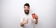 © Vulp - Bearded man holding a red paper cup of coffee or tea and pointing at it on white background in white t-shirt.