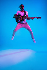 Wall Mural - Young african-american musician playing the guitar in jump like a rockstar on blue studio background in neon light. Concept of music, hobby. Joyful attractive guy improvising. Retro colorful portrait.