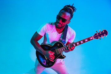 Wall Mural - Young african-american musician playing the guitar like a rockstar on blue studio background in neon light. Concept of music, hobby. Joyful attractive guy improvising. Retro colorful portrait.