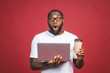 © denis_vermenko - Young surprised african man standing and using laptop computer isolated over red background. Drinking coffee or tea.
