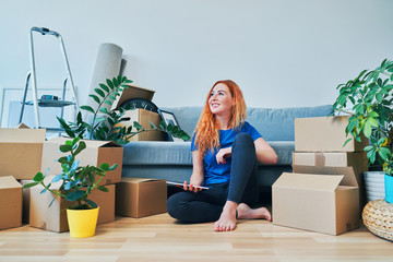 Wall Mural - Cheerful young woman sitting on floor in new apartment and looking away