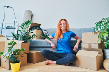 Wall Mural - Portrait of young woman sitting on floor among boxes in new apartment and looking at camera