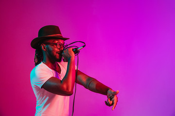 Wall Mural - Young african-american jazz musician with microphone singing a song on purple studio background in trendy neon light. Concept of music, hobby, inspirness. Colorful portrait of joyful attractive artist