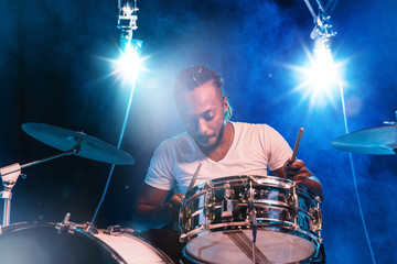 Wall Mural - Young african-american jazz musician or drummer playing drums on blue studio background in glowing smoke around him. Concept of music, hobby, inspirness. Colorful portrait of joyful attractive artist.