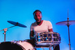 © master1305 - Young african-american jazz musician or drummer playing drums on blue studio background in trendy neon lights. Concept of music, hobby, inspirness. Colorful portrait of joyful attractive artist.