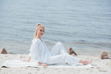 Wall Mural - cheerful blonde young woman sitting on yoga mat near river