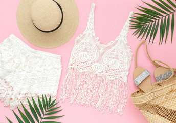 White crochet tank top, shorts, straw boater hat, wicker beach bag, silver glitter flat sandans, tropical palm leaves on pink background. Overhead view of woman's beach outfit. Flat lay, top view.