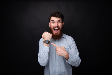 Photo of a handsome frustrated man with beard, delaying and pointing at watch, with shocked face, standing over dark isolated background