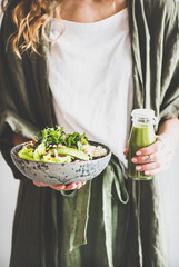 Wall Mural - Healthy dinner or lunch. Woman in linen shirt standing and holding vegan superbowl or Buddha bowl with hummus, vegetable, fresh salad, beans, couscous and avocado and smoothie in hands