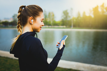 Young smiling girl making sport and running in the park using her phone to listen the music with wireless headphones on sunset in the city watching the screen and having communication talking chat