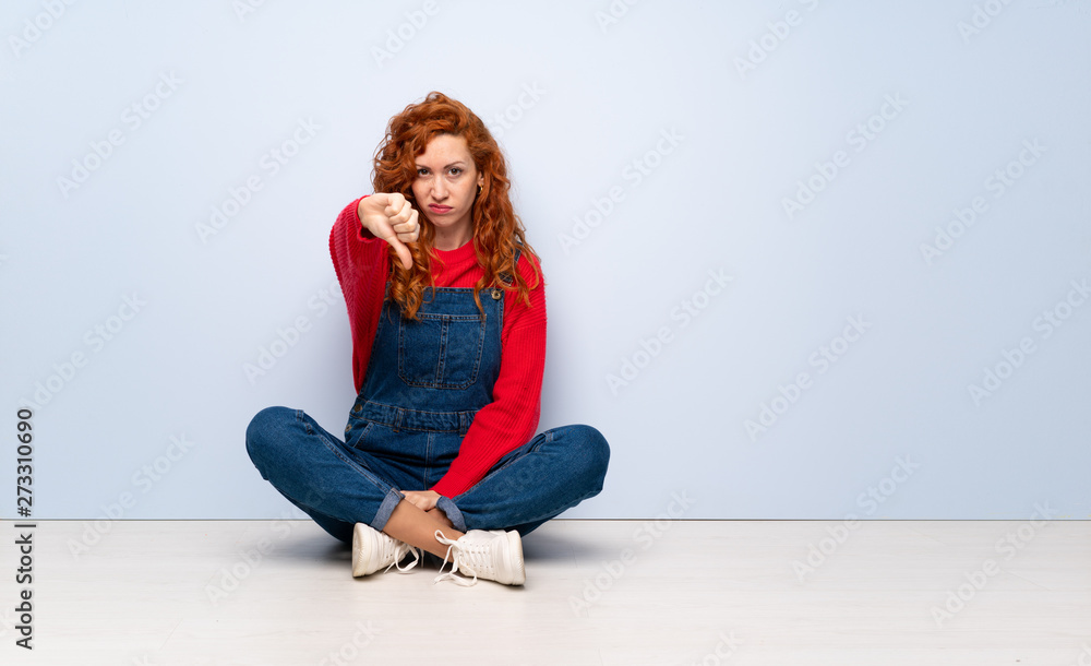 Redhead woman with overalls sitting on the floor showing thumb down ...