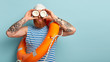 © WHstudio Leushin N - Horizontal shot of ginger male sailor looks into distance, uses coconut as binoculars, wears rubber swimhat, blue and white striped vest, has tattoo, models indoor over blue wall with free space