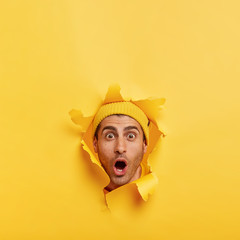 Wall Mural - Vertical shot of stupefied unshaven man stares at camera, keeps head through torn paper hole over yellow background, expresses great disbelief, free space above for your promotional content.