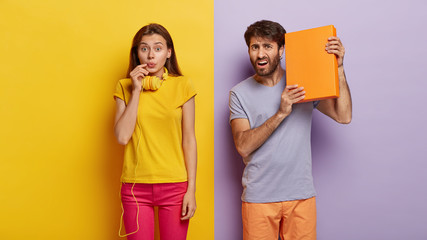 Handsome man holds orange box, cannot understand what is inside, wears casual t shirt and shorts, poses over purple wall. Impressed young female with headphones poses near against yellow background