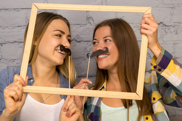 two beautiful girls are laughing, they put a mustache to their face