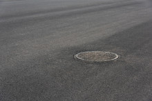 There Is A Metal Manhole Cover On The Wide Black Tarmac Of The City, The Urban Pavement Drainage System