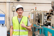 © twinsterphoto - Front view of Asian factory worker with safety hard hat posed looking at camera with happy smile in industrial facilities at heavy industry manufacturing factory. He is giving thumbs up for approval