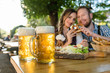 Bier Maßkrüge, Paar sitzt im Biergarten mit Brotzeit im Hintergrund