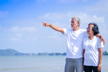 Wall Mural - An elderly Asian couple wears a white shirt. They walked to the beach. He stood hugging each other and pointing his finger at the sea.