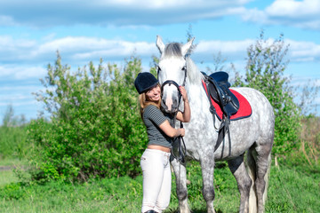 Beautiful cute sexy slim girl jockey with a beautiful smile next to a gray horse in spots in nature on a clear Sunny summer day