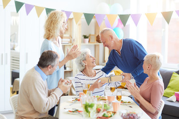 Group of positive excited people celebrating birthday of retired pretty woman at home, handsome man giving gift to birthday woman