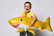 © FAB.1 - Positive little boy hold a balloon in the shape of a yellow shark fish, celebrate the holiday, smiling widely, stand on a light background, are in a good mood