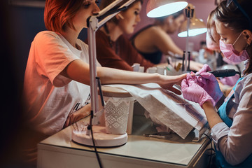 Young attractive manicure master is doing nail treatment for another woman at busy nail studio.