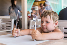 A Little Boy Holds A Knife And Fork And Waits For Food. A Hungry Child Is Sitting At A Table In A Summer Restaurant Waiting For Meals.