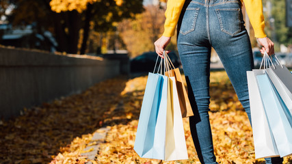 Woman style and fashion. Cropped back view of lady in jeans standing with shopping bags. Blur fall city background. Copy space.