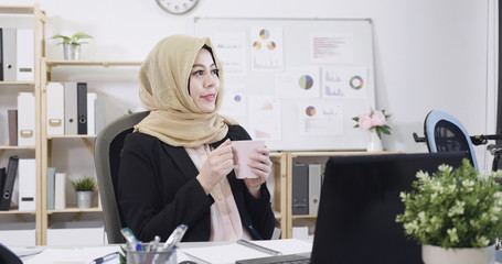 Wall Mural - Attractive young arabic businesswoman sitting at workplace desk in office relax having break. malay lady drinking coffee holding tea cup and smiling looking aside with joy emotion. copy space.