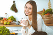 © rogerphoto - Young happy woman is holding white cup and looking at the camera while sitting at wooden table in the kitchen among green vegetables