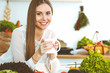 © rogerphoto - Young happy woman is holding white cup and looking at the camera while sitting at wooden table in the kitchen among green vegetables