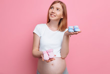 Beautiful Pensive Pregnant Woman Holding Blue And Pink Baby Booties, Posing In The Studio On A Pink Background