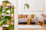 Fototapeta Boho - The stylish boho interior of living room in nice apartment with gray sofa, honey yellow plaid and pillows, a lot of plants and elegant accessories. Handmade macrame shelf planter hanger and paintings.