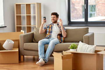 moving, people and real estate concept - happy man with smartphone and boxes sitting on sofa and having video call at new home