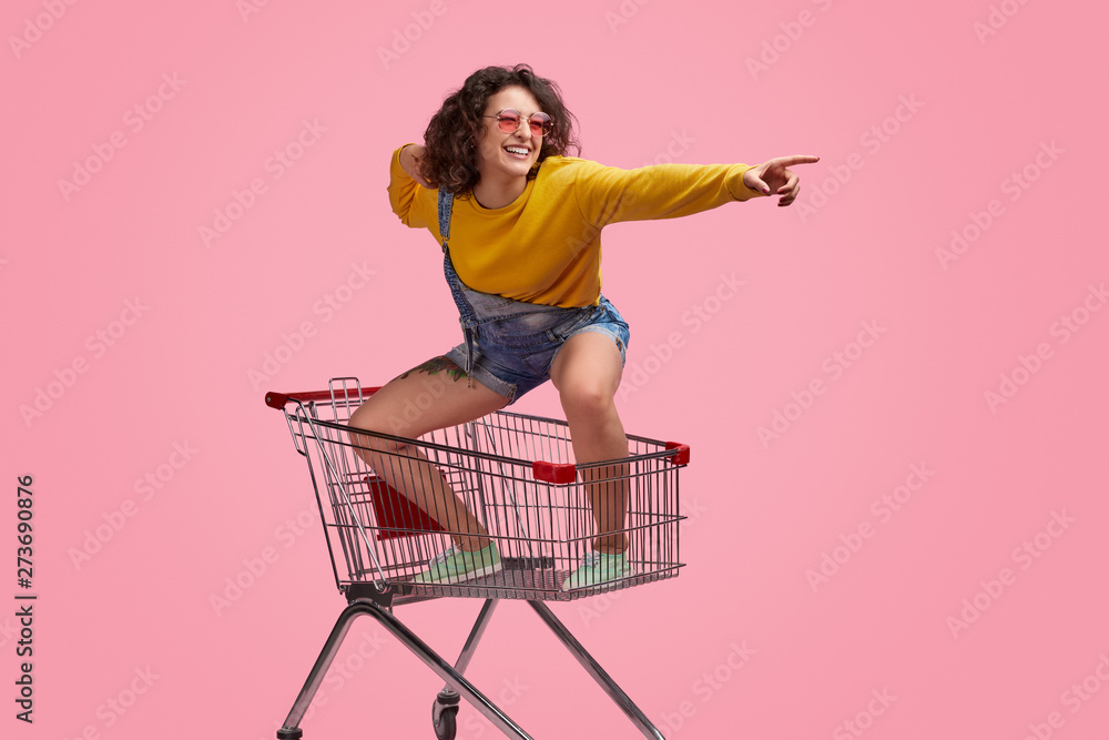 Cheerful young woman riding forward on shopping cart - obrazy, fototapety, plakaty 