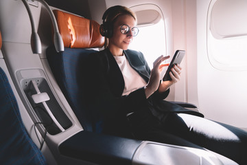 Wall Mural - Young caucasian woman sitting near window with smartphone connected to wifi internet during flight on board, female passenger in headphones for noise cancellation search songs in online shop on app