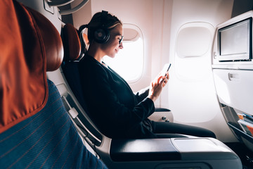 Wall Mural - Side view of woman in formal wear watching online positive video while using wireless internet connection on board, female passenger reading news while listening audio record via headphones