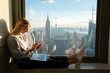 © BullRun - Trendy caucasian female texting message on smartphone while sitting on office window sill with laptop computer. Young student hipster blogger using application on mobile phone.Exchange abroad New York