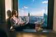 © BullRun - Blonde hair caucasian female student closed her eyes while enjoying warm evening sunset light though hotel window with panoramic New York city views. Hipster girl traveler working on laptop computer