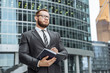 © Artem - Portrait of a successful business man in a suit and glasses with a notebook in his hands on the background of the business center