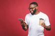 © denis_vermenko - Typing a message. Cheerful black man typing something on the mobile phone, drinking coffee and smiling while standing isolated against red background.