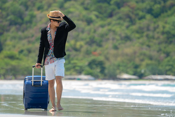 Young asian businessman traveler in casual suit walking barefoot with blue suitcase luggage on the island beach in summer day holidays vacation travel with blurred island, beach and sea backgrounds